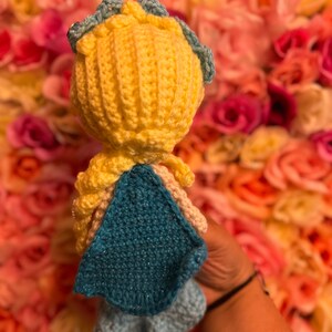 May include: A crocheted doll with yellow hair, a blue crown, and a blue cape. The doll is wearing a blue dress and is standing in front of a pink and white flower wall.