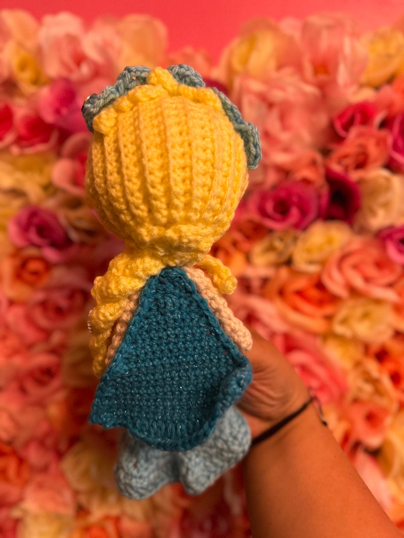 May include: A crocheted doll with yellow hair, a blue crown, and a blue cape. The doll is wearing a blue dress and is standing in front of a pink and white flower wall.