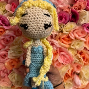 May include: A crocheted doll with blonde hair, a blue dress, and a crown. The doll is holding a long braid of yarn in its right hand. The doll is posed against a background of pink and white artificial flowers.