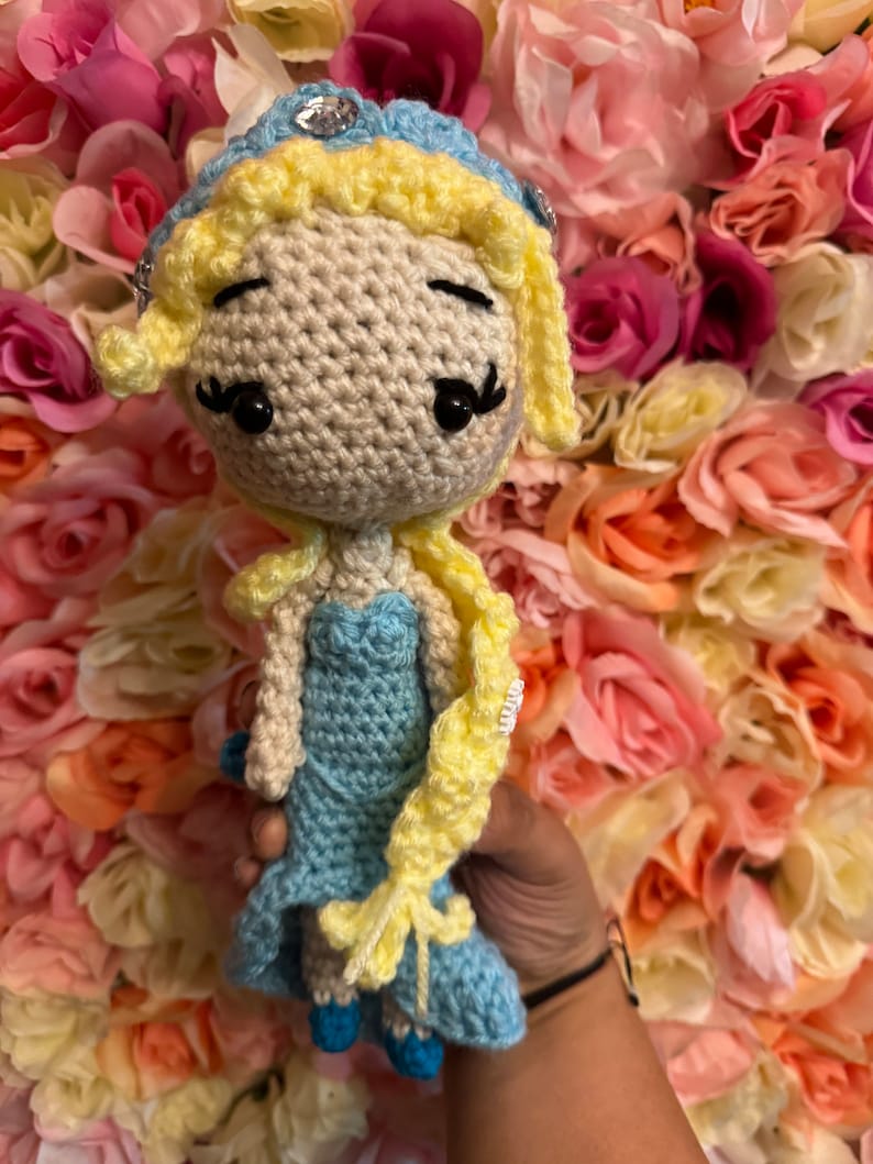 May include: A crocheted doll with blonde hair, a blue dress, and a crown. The doll is holding a long braid of yarn in its right hand. The doll is posed against a background of pink and white artificial flowers.