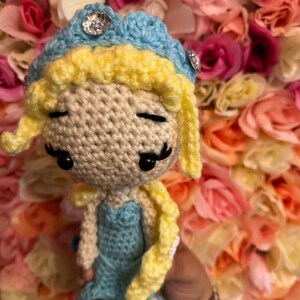 May include: A crocheted doll with blonde hair, a blue dress, and a blue and yellow crown. The doll has black eyes and a sweet expression.