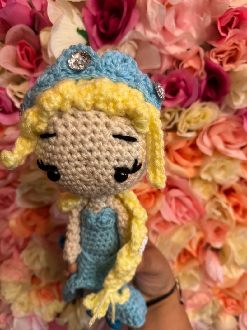 May include: A crocheted doll with blonde hair, a blue dress, and a blue and yellow crown. The doll has black eyes and a sweet expression.
