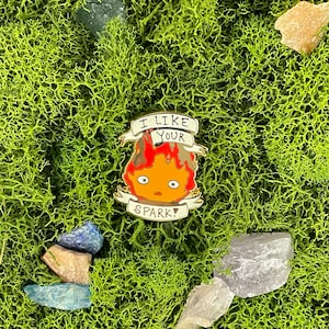 May include: A colorful enamel pin featuring a cartoon fire demon with the text "I LIKE YOUR SPARK!". The pin has a red and orange flame design with a white banner. The pin is set against a background of green moss and crystals.