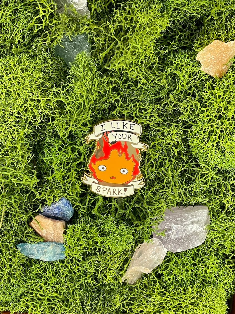 May include: A colorful enamel pin featuring a cartoon fire demon with the text "I LIKE YOUR SPARK!". The pin has a red and orange flame design with a white banner. The pin is set against a background of green moss and crystals.