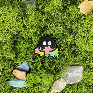 May include: A black cartoon character with a floral design is set against a backdrop of vibrant green moss and various crystals. The character has a round body and two white eyes, with a colorful flower design.