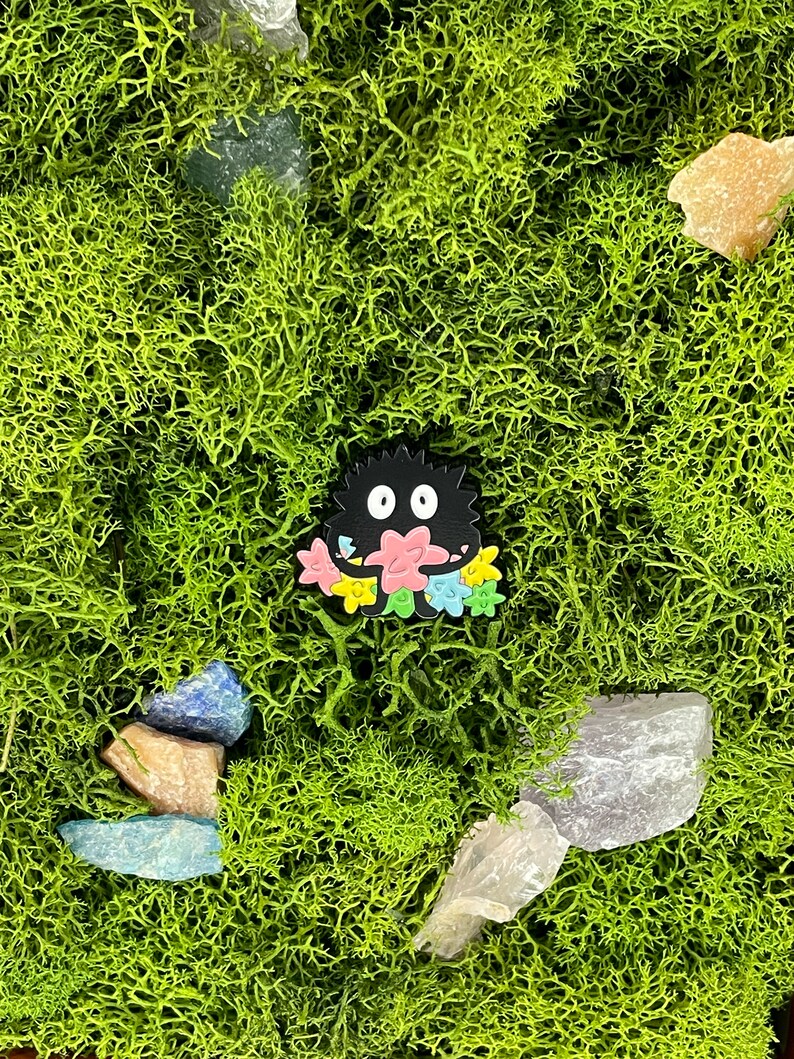 May include: A black cartoon character with a floral design is set against a backdrop of vibrant green moss and various crystals. The character has a round body and two white eyes, with a colorful flower design.