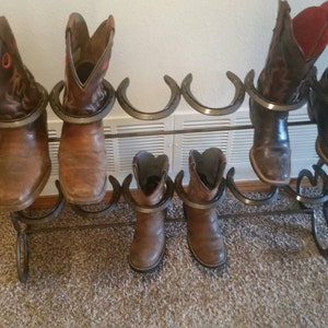 May include: A metal boot rack with six horseshoe-shaped holders. The rack is holding six pairs of brown and black cowboy boots.
