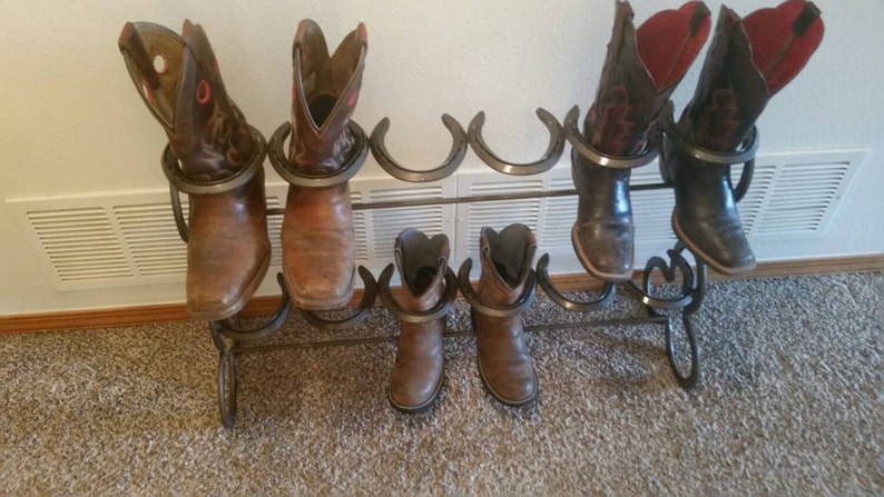 May include: A metal boot rack with six horseshoe-shaped holders. The rack is holding six pairs of brown and black cowboy boots.