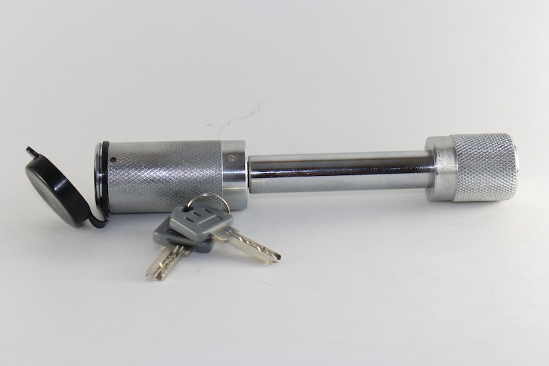 May include: A silver hitch lock with a black cap and two keys. The lock has a threaded end and a smooth, cylindrical body.