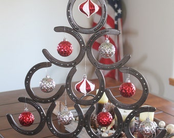 May include: A metal Christmas tree made of horseshoes decorated with red and silver ornaments. The tree is on a wooden table with a wooden tray of pine cones and other decorations.