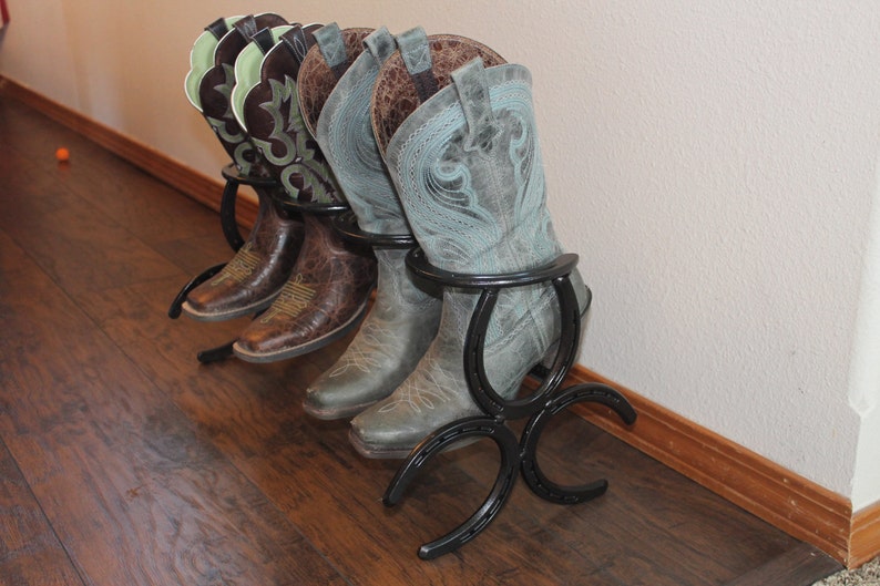 Puede incluir: Un conjunto de tres botas de vaquero se muestran en un perchero de botas de metal negro hecho de herraduras. Las botas son marrones y azules con dise&ntilde;os intrincados.