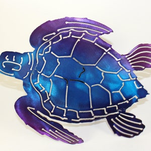 May include: A metal sea turtle wall art sculpture with a blue and purple ombre finish. The turtle is in a swimming position with its head and flippers extended.