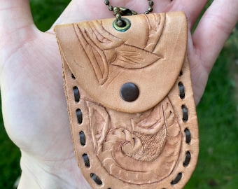 May include: A small, light brown leather keychain with a decorative floral design. The keychain has a metal loop and a flap with a snap closure.