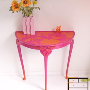 May include: A pink half-round table with orange floral stenciling and a pink ombre finish on the legs. The table is decorated with a vase of orange flowers and a small orange deer figurine.