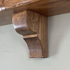 May include: A rustic, wooden mantelpiece with a natural finish. The mantel is supported by a decorative bracket. The wood grain is visible, and the piece has a warm, inviting tone. The mantel is designed for mounting on a wall.