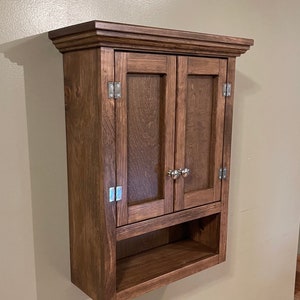 May include: A wall-mounted wooden cabinet with two doors and a small shelf below. The cabinet is made of dark brown wood and has silver-colored hinges and door knobs. The cabinet is mounted on a light brown wall.