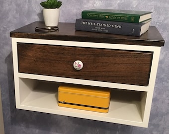 May include: A floating nightstand with a dark brown top, white frame, and a drawer with a floral knob. The drawer is a medium brown color. A small succulent in a white pot, books, and a yellow tin are on the nightstand.