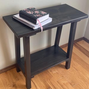 May include: A dark stained wooden side table with a lower shelf. A floral-covered book and a spiral notebook with a pink ribbon are on the tabletop. The table is approximately 24 inches tall.