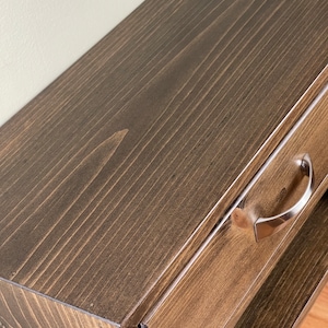 May include: Close-up of a stained wooden bedside table with a drawer and a lower shelf. The wood has a dark brown finish with visible grain. A silver-coloured handle is attached to the drawer. The bedside table is a piece of bedroom furniture.