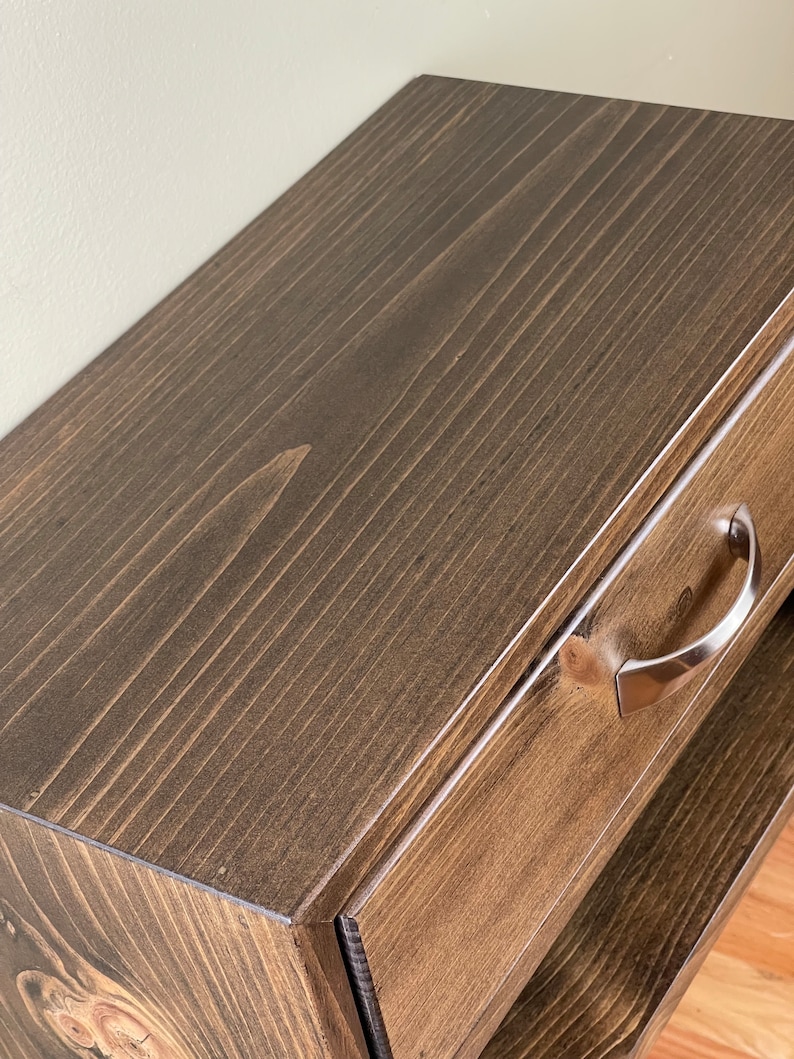 May include: Close-up of a stained wooden bedside table with a drawer and a lower shelf. The wood has a dark brown finish with visible grain. A silver-coloured handle is attached to the drawer. The bedside table is a piece of bedroom furniture.