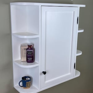 May include: White wall-mounted cabinet with a single door and open shelving. The cabinet has a black knob and hinges. A blue mug, a bottle of lotion, and a box are on the shelves. The cabinet is designed for bathroom storage.