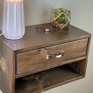 May include: A dark stained wooden bedside table with a drawer and open shelf. A silver handle is on the drawer. A white lamp with a grey lampshade and a geometric terrarium with a plant are on top.