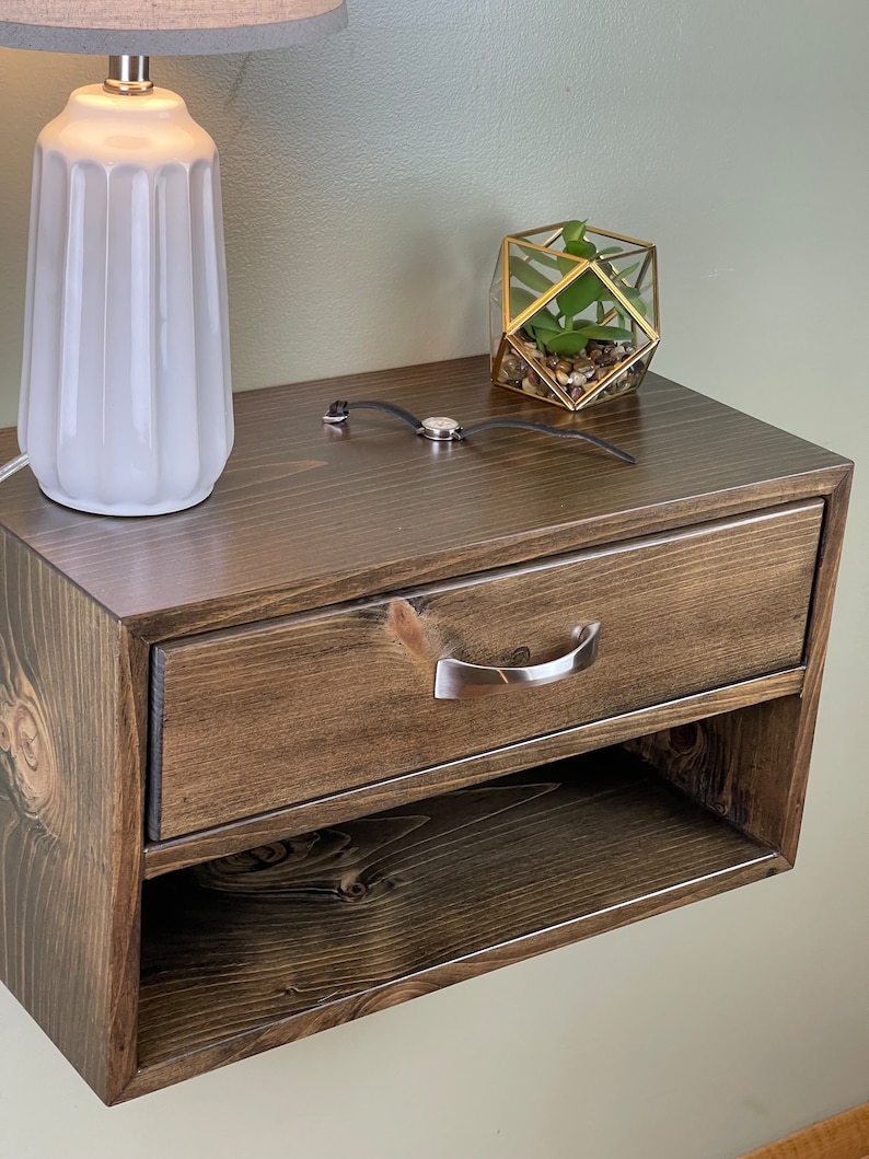 May include: A dark stained wooden bedside table with a drawer and open shelf. A silver handle is on the drawer. A white lamp with a grey lampshade and a geometric terrarium with a plant are on top.