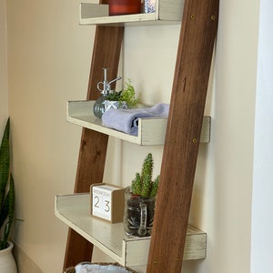 Puede incluir: Estanter&iacute;a de escalera de madera r&uacute;stica con cuatro estantes. Los estantes pintados de blanco con bordes desgastados exhiben accesorios de ba&ntilde;o, como una vela, plantas, toallas y un calendario que indica mi&eacute;rcoles 23 de noviembre.