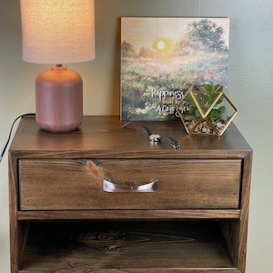 May include: A dark stained wooden floating bedside table with a drawer and open shelf. A small lamp with a beige shade and a decorative picture frame with the text "happiness trials keep courage" sit on top. A small terrarium and watch are also visible.