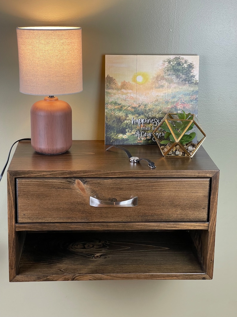 May include: A dark stained wooden floating bedside table with a drawer and open shelf. A small lamp with a beige shade and a decorative picture frame with the text "happiness trials keep courage" sit on top. A small terrarium and watch are also visible.