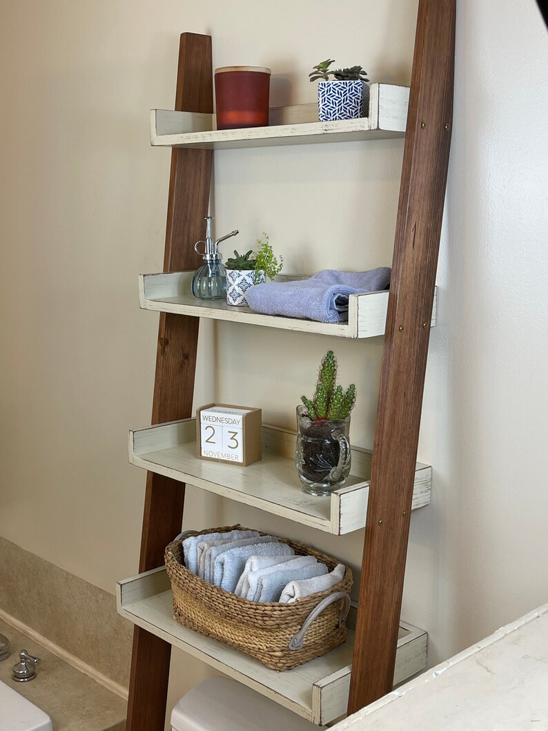 Puede incluir: Una estanter&iacute;a de escalera de madera con cuatro estantes, que muestra accesorios de ba&ntilde;o. Los estantes contienen una vela, plantas en macetas, un pulverizador, una toalla enrollada, un calendario de madera, un cactus y una cesta de toallas dobladas.