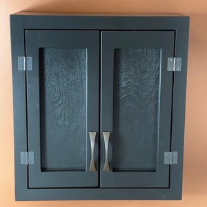 May include: A black wooden cabinet with two doors and silver handles. The cabinet has a simple design and is mounted on a wall.