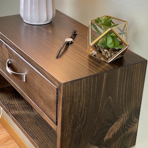 May include: A dark stained wooden bedside table with a drawer and open shelf. A white ceramic vase, a wristwatch, and a geometric glass terrarium with a succulent plant are on top. The table has a brushed metal drawer pull.