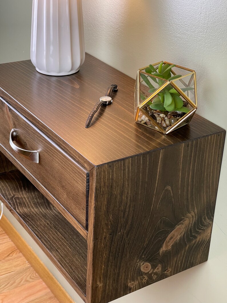 May include: A dark stained wooden bedside table with a drawer and open shelf. A white ceramic vase, a wristwatch, and a geometric glass terrarium with a succulent plant are on top. The table has a brushed metal drawer pull.