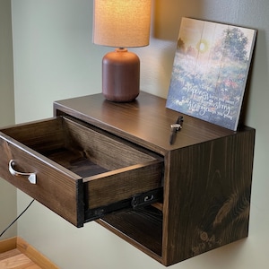 May include: A dark brown floating nightstand with an open drawer, a silver handle, and a small lamp. A framed print with text and a small key are on top. The nightstand is mounted on a light green wall.