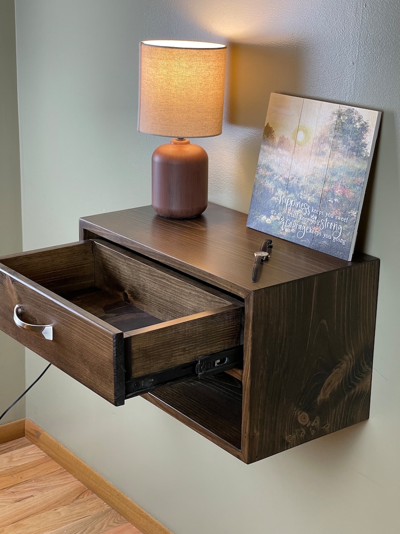 May include: A dark brown floating bedside table with an open drawer, a silver handle, and a small lamp. A framed print with text and a small key are on top. The table is mounted on a light green wall.