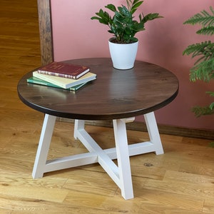 May include: A round wooden coffee table with a dark brown top and white painted legs. The table has a cross-shaped support in the middle of the legs. A potted plant and three books are on the table.