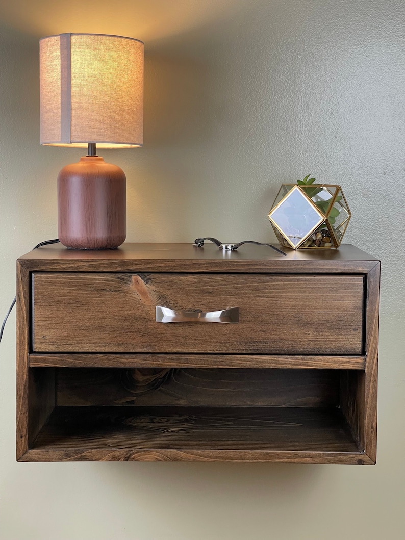 May include: A dark brown, floating bedside table with a drawer and open shelf. A brown lamp with a beige shade sits on top, along with a geometric glass terrarium. The table has a silver-toned drawer pull.