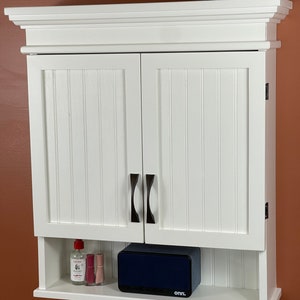 May include: White wall mounted cabinet with two doors and a shelf. The cabinet has a decorative trim around the top and bottom. The doors have vertical grooves and black handles. The shelf has a small bottle of liquid and two small bottles of nail polish on it. A black speaker with the text "onn." is on the shelf.