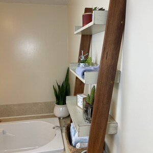 Puede incluir: Estanter&iacute;a de escalera de madera con cuatro estantes, pintada de blanco con un acabado envejecido. La escalera tiene soportes de madera marr&oacute;n oscuro. Los estantes est&aacute;n decorados con plantas, toallas y una cesta de mimbre. La estanter&iacute;a est&aacute; apoyada contra una pared beige en un ba&ntilde;o.