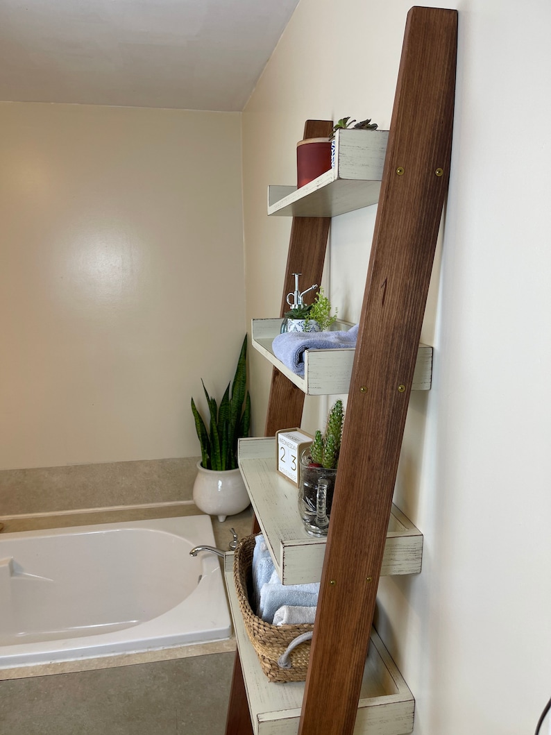Puede incluir: Estanter&iacute;a de escalera de madera con cuatro estantes, pintada de blanco con un acabado envejecido. La escalera tiene soportes de madera marr&oacute;n oscuro. Los estantes est&aacute;n decorados con plantas, toallas y una cesta de mimbre. La estanter&iacute;a est&aacute; apoyada contra una pared beige en un ba&ntilde;o.