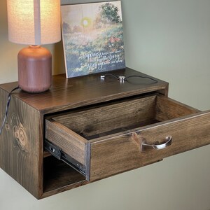 May include: A dark stained wooden floating bedside table with an open drawer. A small lamp with a beige shade and a wooden base sits on top. A framed picture with the text "happiness keeps you sweet" is also on top.
