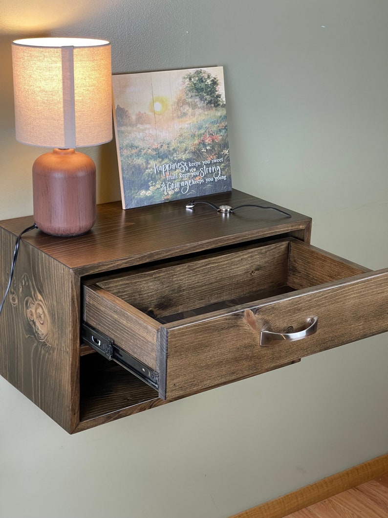 May include: A dark stained wooden floating bedside table with an open drawer. A small lamp with a beige shade and a wooden base sits on top. A framed picture with the text "happiness keeps you sweet" is also on top.
