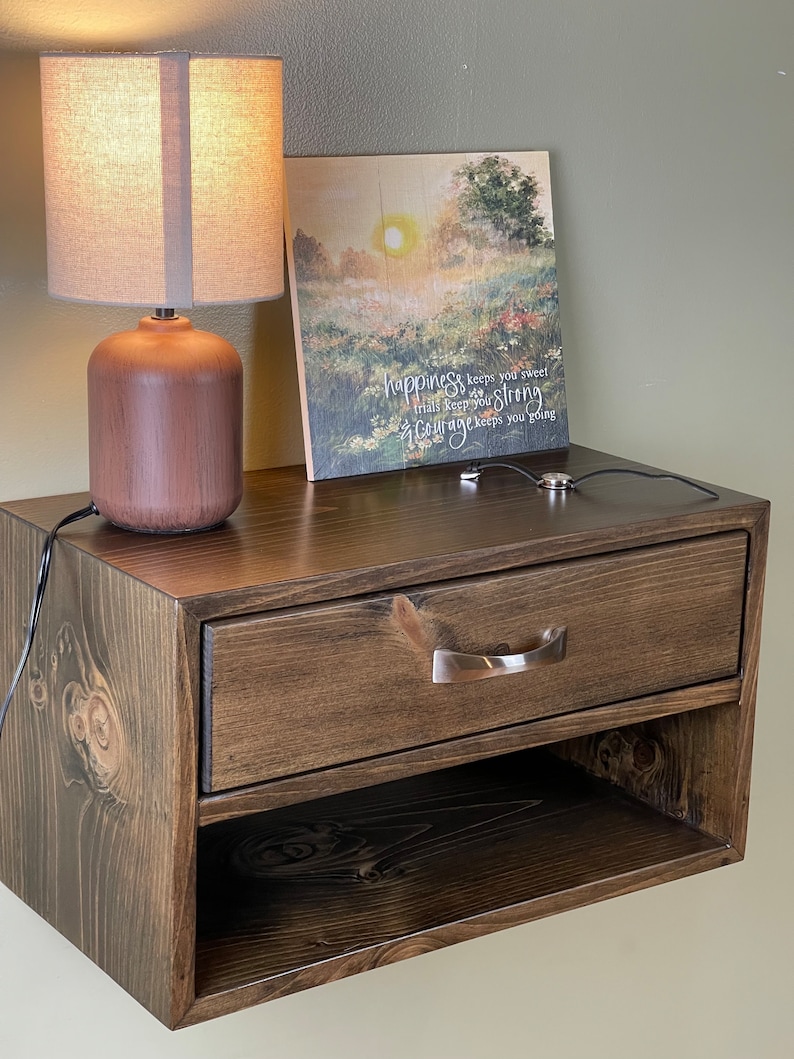 May include: A dark brown wooden floating nightstand with a drawer and open shelf. A brown lamp with a beige shade sits on top, next to a framed landscape print with the text "happiness keeps you sweet, trials keep you strong, courage keeps you going."