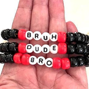 May include: Three beaded bracelets with the words "BRUH", "DUDE", and "BRO" spelled out in white letter beads. The bracelets have black and red beads. The bracelets are held in a hand.