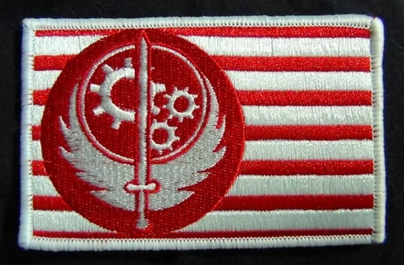 以下が含まれることがあります： 白と赤の刺繍パッチ。円の中に剣と歯車の白いシンボルが描かれている。シンボルは白のストライプが入った赤い背景に描かれている