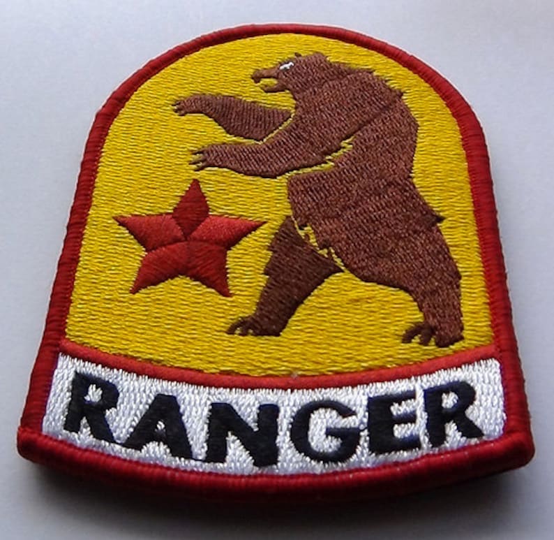 May include: A yellow and red embroidered patch with a brown bear standing on its hind legs. A red star is to the left of the bear. The text "RANGER" is in black letters on a white background at the bottom of the patch.