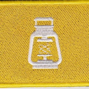 Puede incluir: Un parche de tela amarillo con una imagen bordada en blanco de una linterna.
