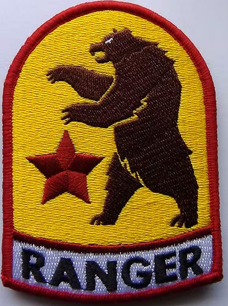 May include: A yellow and red embroidered patch with a brown bear standing on its hind legs. The bear is facing left and has a red star in front of it. The patch has the word "RANGER" in black letters on a white background at the bottom.