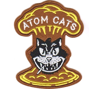 May include: A brown and yellow embroidered patch with a cartoon cat's face and the words "ATOM CATS" in a white font.